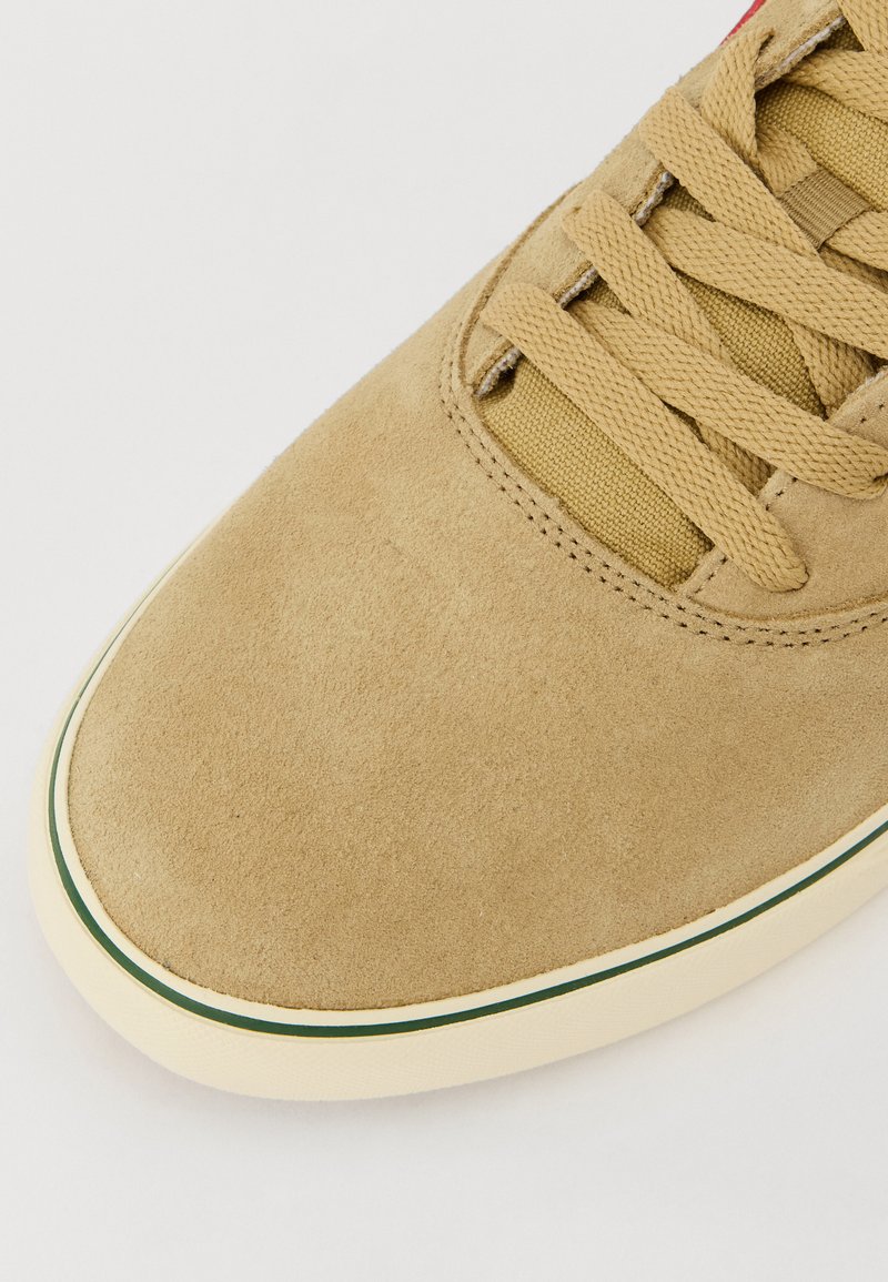 Zapatilla de ante marrón claro con puntera redondeada, superficie texturizada, cordones beige y una suela media blanca acentuada con una franja verde.