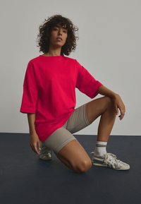 T-shirt oversize rossa in cotone, pantaloni corti beige a coste lunghi fino al ginocchio e sneaker grigie con accenti argentati. Il modello si posa su un ginocchio.