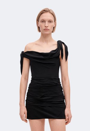 Un mannequin se tient devant un fond uni, vêtu d’une mini-robe noire drapée, épaules dénudées et à jupe froncée.