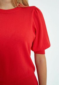 Sarkana adīta krekla augšdaļa ar apaļu kakla izgriezumu, īsām pūkainām piedurknēm un ribotu apakšmalu. Materiāls izskatās mīksts ar gludu tekstūru.
