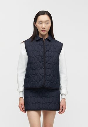 Vrouw die een marineblauw doorgestikt vest en een rok draagt over een wit shirt met lange mouwen, staand tegen een effen lichte achtergrond.