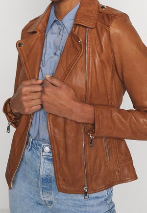 Persoon die een bruine leren jas aanpast over een blauw overhemd met knopen en lichtblauwe jeans, met zichtbare ritsdetails en textuur van de jas.