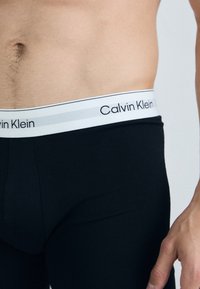 Črno spodnje perilo Calvin Klein z belim pasom, na katerem je ime blagovne znamke in sive poudarjene črte. Povečani posnetek teksture tkanine.