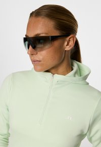 Felpa con zip verde chiaro in materiale morbido con logo bianco. Occhiali da sole neri con grandi lenti scure e montatura spessa.