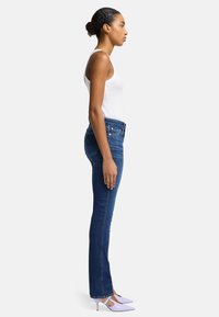 Jeans en denim bleu skinny avec une taille mi-haute, associés à un débardeur blanc et des chaussures à talons lavande, présentés en vue de profil.