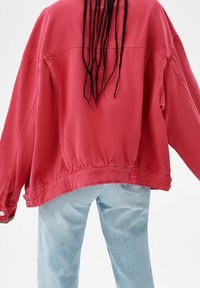 Przesadnie duża czerwona dżinsowa kurtka o teksturowanym wykończeniu, widoczne szwy oraz zapięcia na guziki przy mankietach, zestawiona z jasnoniebieskimi dżinsami.