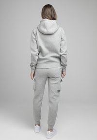 Persona vestida con un hoodie gris claro y pantalones de chándal cargo a juego, de pie de espaldas a la cámara sobre un fondo gris liso.