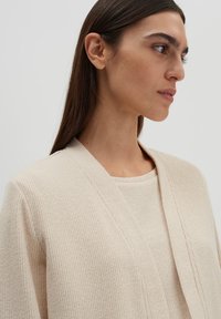Beige strukturierter Cardigan mit V-Ausschnitt und offener Front. Das Material wirkt weich und gestrickt und zeigt ein dezentes Rippenmuster.
