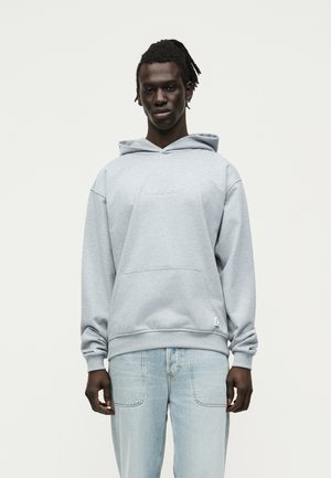 Ung mand med mørk hud og dreadlocks iført en lys grå hættetrøje og lyseblå jeans, stående foran en ensfarvet hvid baggrund.