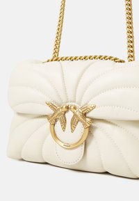 Grietos odos rankinė su siuvinėtu raštu, turinti auksinę grandinėlę ir užsegimą su dviem paukščiais. Matomas išskirtinis logotipo detalė.