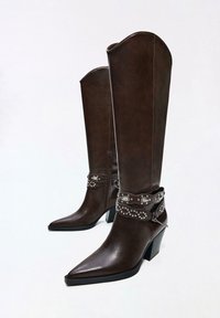 Ciemnobrązowe skórzane kozaki za kolano z zaostrzonymi noskami, słupkowymi obcasami i ozdobnymi paskami z ćwiekami oraz metalowymi akcentami wokół kostek.