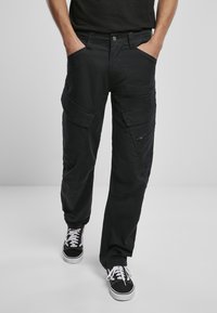 Schwarze Cargo-Hosen aus Baumwollmaterial. Verfügt über mehrere Taschen, eine lockere Passform und Reißverschluss-Akzente. Kombiniert mit schwarz-weißen Sneakers.