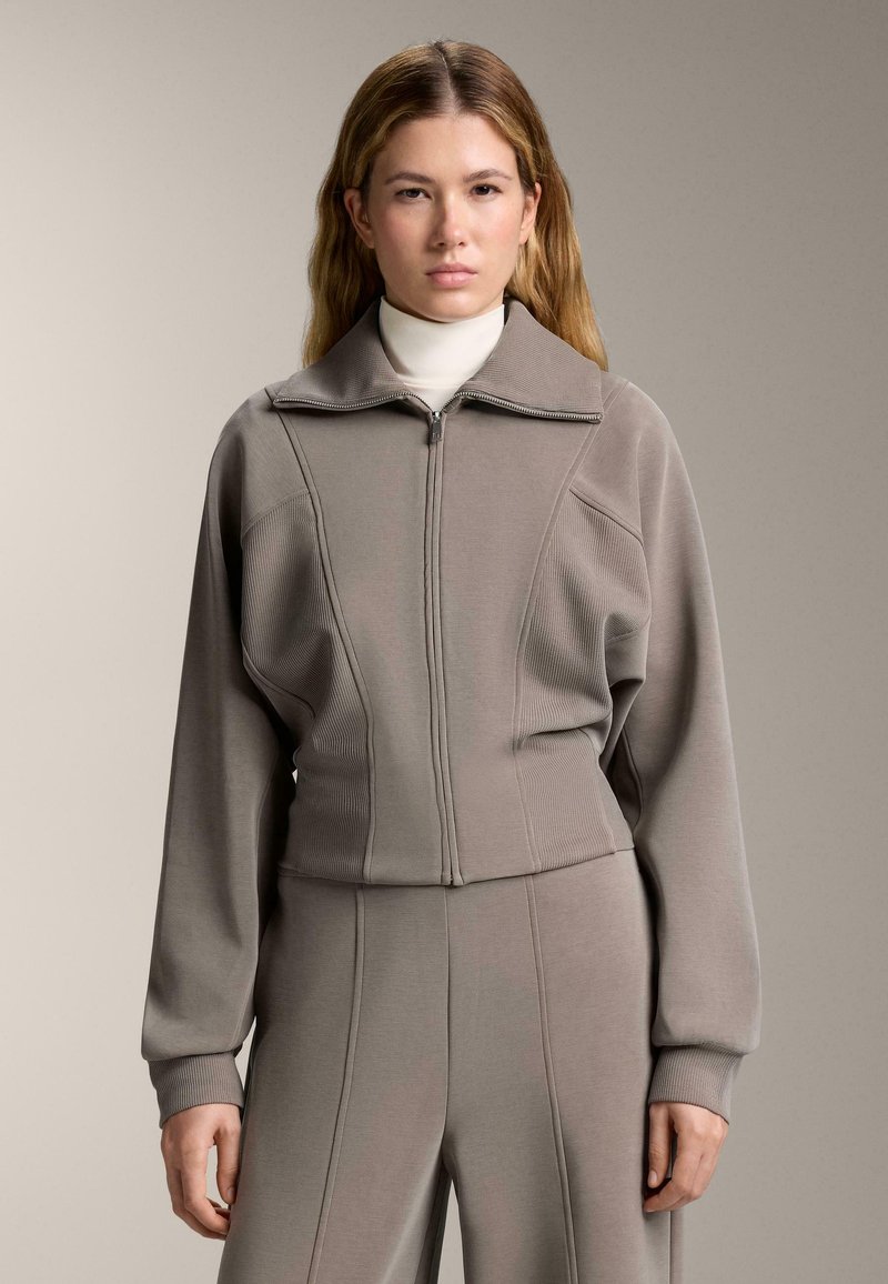 Svijetlosivi dukserica s patentnim zatvaračem, širokim ovratnikom i rebrastim detaljima, u kombinaciji s odgovarajućim hlačama. Mekana, teksturirana tkanina s uskim krojem.