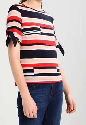 Kobieta w krótkim, paskowanym topie z kokardkami na rękawach, w połączeniu z niebieskimi dżinsami, stoi na jednolitym tle.