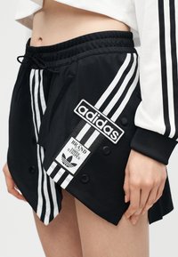 Μαύρη φούστα Adidas με τρεις λευκές ρίγες, ετικέτες με λογότυπα και κουμπιά snap, συνδυασμένη με λευκό μακρυμάνικο τοπ που διαθέτει μαύρες ρίγες.