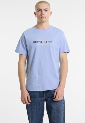 Svijetloplavi pamučni majica kratkih rukava, s logom "GUESS JEANS" u crnom tekstu centriranim sprijeda, u kombinaciji sa plavim trapericama.