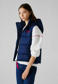 Ναυτικό μπλε γιλέκο με πλούσια σχεδίαση, κουκούλα και κόκκινες λεπτομέρειες. Φορεμένο πάνω από ένα λευκό πουκάμισο, σε συνδυασμό με μπλε τζιν.