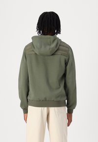 Olivengrøn hættetrøje med tekstureret, polstret design på skuldrene, elastiske manchetter og kant; blødt stof. Model iført lyse beige bukser.