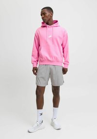 Sudadera con capucha rosa y bolsillo delantero, combinada con pantalones cortos grises. Logo blanco en el pecho, complementado por zapatillas blancas y calcetines con detalles en negro.