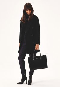 Black leather tote bag with dual handles, smooth texture, and a logo. Worn with a black coat and high-heeled ankle boots, styled with tailored pants.