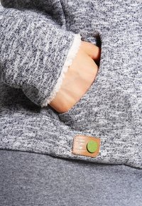 Harmaa neulepaita mukavalla rakenteella, jossa on sivutasku ja nahkainen merkki vihreällä korostuksella. Hihansuut vuorattu pehmeällä materiaalilla.