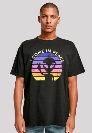 Black cotton t-shirt featuring a graphic of an alien in a peace sign against a circular sunset gradient of pink, purple, and yellow stripes.