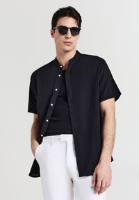 Schwarzes kurzärmeliges Hemd mit Stehkragen, aus strukturiertem Stoff und weißen Knöpfen, getragen über einem schwarzen Tanktop und weißen Hosen.