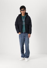 Sort puffjakke med hætte, grøn sweatshirt med tekst, blå jeans og hvide sneakers; bærer sorte solbriller. Står foran en lys baggrund.