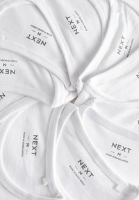 Witte katoenen tanktops in een cirkelvormige opstelling, met bedrukte labels met de tekst "NEXT M Made in Bangladesh." Eenvoudig ontwerp, gladde textuur.
