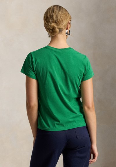 Žena s blond vlasmi v nízkom drdole, ktorá nosí strieborné kruhové náušnice, zelené tričko s krátkym rukávom a tmavé nohavice, otočená chrbtom k neutrálnemu pozadiu.