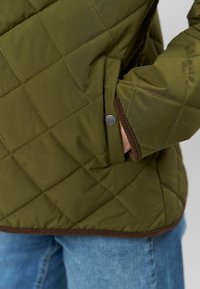 Prešívaná olivovo zelená bunda s vreckom na patent a hnedým lemovaním pozdĺž spodného okraja a manžiet. Hladká, zateplená textúra látky.