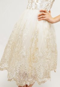 Robe blanche avec un motif floral doré brodé, jupe en tulle et ourlet en forme de vague. Le corsage présente une texture satinée et une coupe ajustée.