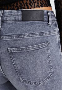 Vedere din spate a unei perechi de blugi gri din denim cu o etichetă neagră din piele cu marca "NOISY MAY", purtați cu un top negru cu model.