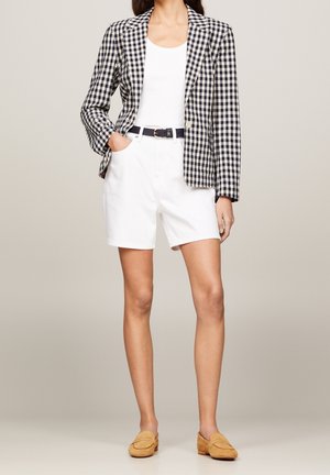 Kvinde iført sort-hvid ternet blazer, hvid top, hvide shorts med sort bælte og lysebrune loafers, stående med den ene hånd i lommen.