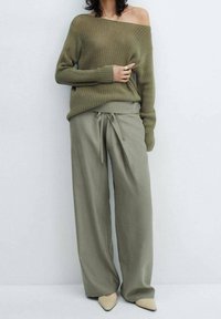 Overmaatse olijfgroene gebreide trui met een off-shoulder ontwerp, gecombineerd met ruime bijpassende broeken en puntige beige schoenen.