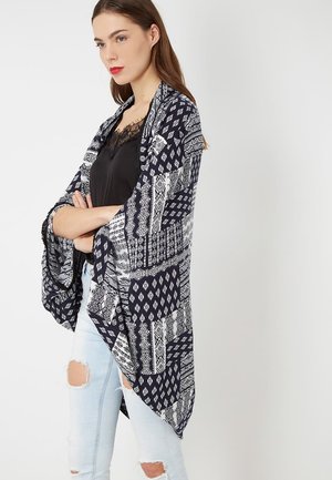 Joven con los brazos cruzados, vestida con un top negro de encaje, un chal azul y blanco de patrón patchwork y jeans azules claros rasgados, mirando hacia la izquierda.