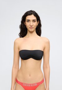 Γυναίκα με μακριά σκούρα μαλλιά που φοράει μαύρο στράπλες σουτιέν και κοραλί εσώρουχα Calvin Klein, στέκεται μπροστά σε απλό ανοιχτό φόντο.