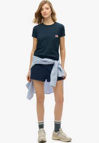 Marineblauwe katoenen t-shirt met een borstzak, gecombineerd met marineblauwe shorts. Blauw-wit gestreept shirt om de taille geknoopt. Lichtgekleurde sneakers.
