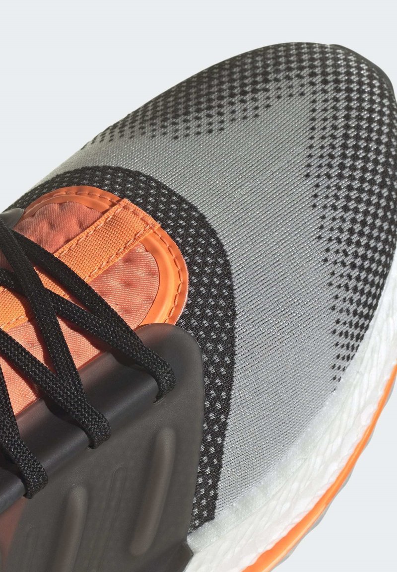 Zapatilla deportiva con un upper textil gris y acentos en negro y naranja. Patrones texturizados y cordones negros sobre una mediasuela blanca.