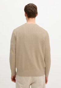 Homme debout de dos, portant un pull beige côtelé à manches longues et un pantalon beige clair, sur un fond blanc uni.