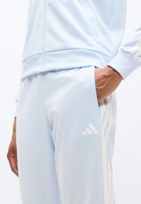 Αθλητική φόρμα ανοιχτού γαλάζιου χρώματος με ζακέτα με φερμουάρ και στενά παντελόνια με λευκές τρεις ρίγες και ανάγλυφο λογότυπο στα παντελόνια.