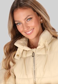 Lys beige pufjakke med høj krave, lynlås lukning og guldfarvet hardware detaljer. Glat tekstur med quiltet design.