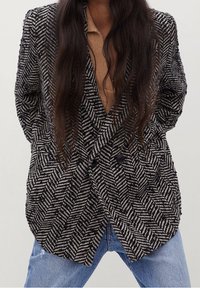 Dwurzędowa marynarka w czarno-białą jodełkę z wyciętymi klapami, czarnymi guzikami i teksturowanym materiałem, połączona z jasnoniebieskimi dżinsami.