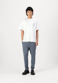 Camiseta blanca de algodón con un gráfico azul en el lado izquierdo del pecho, combinada con pantalones ajustados de color gris claro y zapatos negros. Diseño simple, aspecto casual.