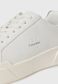 Beli športni čevlji z gladkim usnjenim zgornjim delom, zaobljenim nosom, toniranimi vezalkami in povišanim gumijastim podplatom. Imajo subtilno embosirano blagovno znamko Calvin Klein.