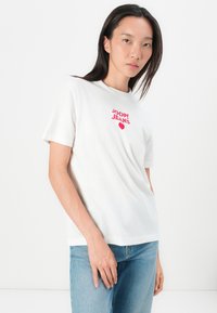 White cotton t-shirt featuring a red printed logo "JOOP! JEANS" with a heart design. Short sleeves and a relaxed fit. Paired with light blue jeans.