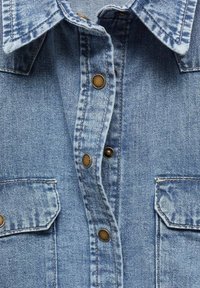 Chemise en denim bleu clair avec col classique, devant boutonné, deux poches poitrine et détails de boutons métalliques marron.