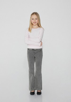 Helles pinkes Langarm-Pullover, graue Schlagjeans, schwarze Ballerinas. Mädchen steht mit verschränkten Armen, glattem Haar, heller Hintergrund.