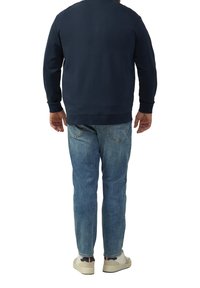 Marineblaue Sweatshirt, Rundhalsausschnitt, gerippte Ärmelbündchen, kombiniert mit hellblauen Jeans. Die Schuhe sind kremfarbig mit schwarzen und roten Akzenten.