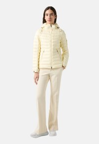 Light yellow puffer jacket with a hood, zippered front, and vertical quilted pattern. Paired with cream-colored pants and white sneakers.